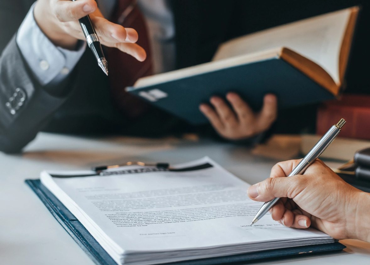 A person is writing on paper with another person holding a book.