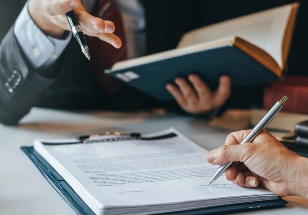 A person is writing on paper with another person holding a book.